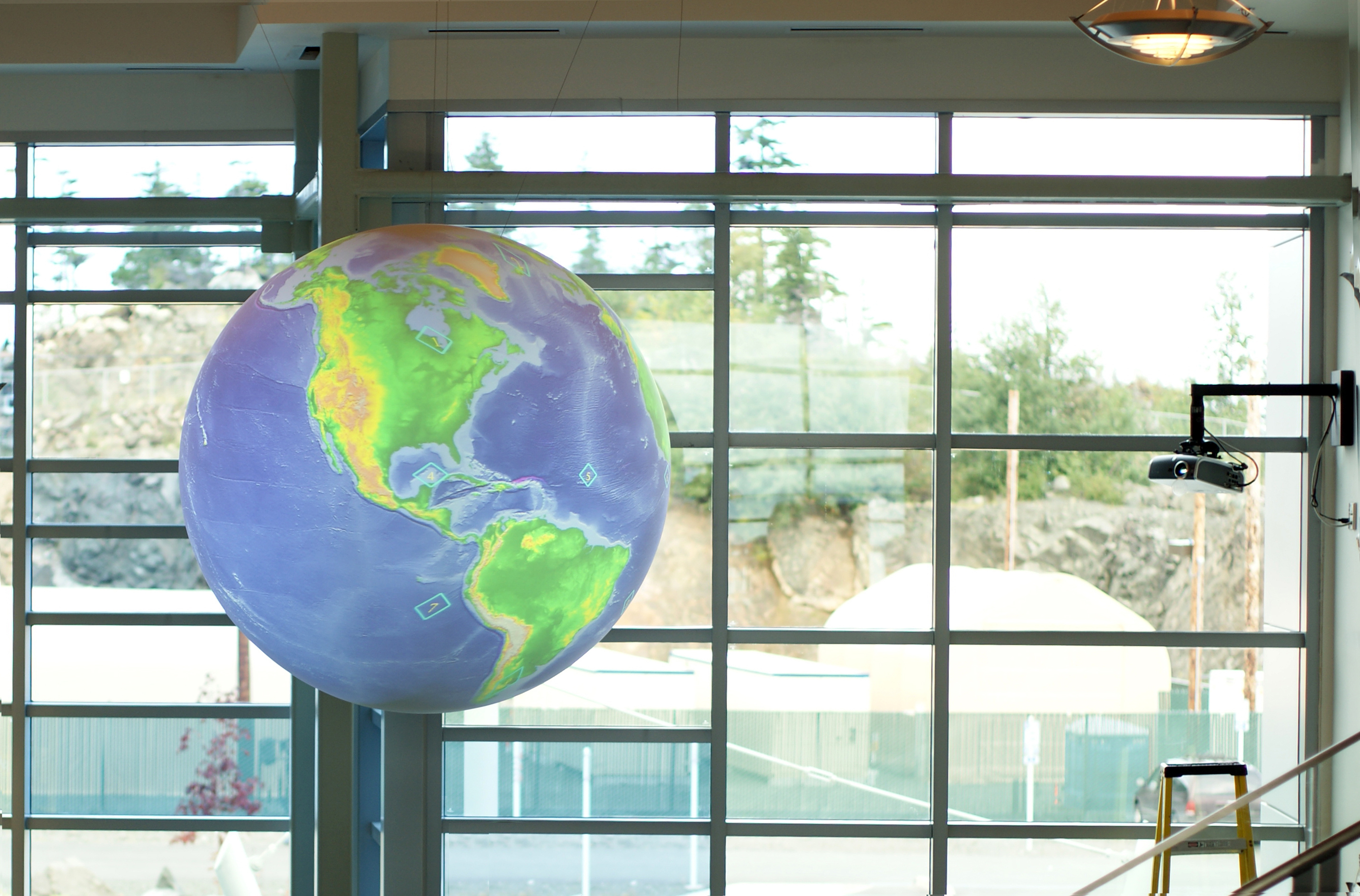 Science On a Sphere hangs in a well-lit room in front of several large windows. Several large tanks can be seen through the window, along with a rock face and some trees