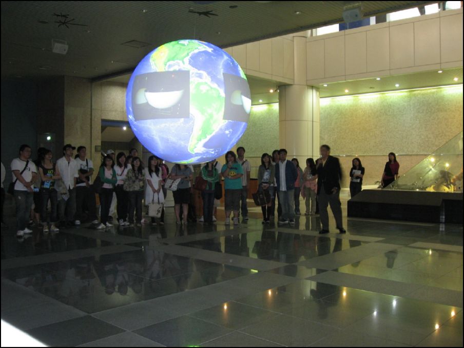 A group of people stand behind Science On a Sphere watching a presentation in a large, well-lit exhibit hall