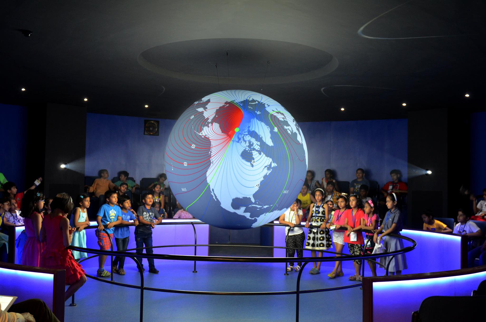 A group of children stand around a railing watching Science On a Sphere in a darkened theater, more children are seated in chairs behind them. Science On a Sphere displays red and blue lines indicating Earth's magnetic declination