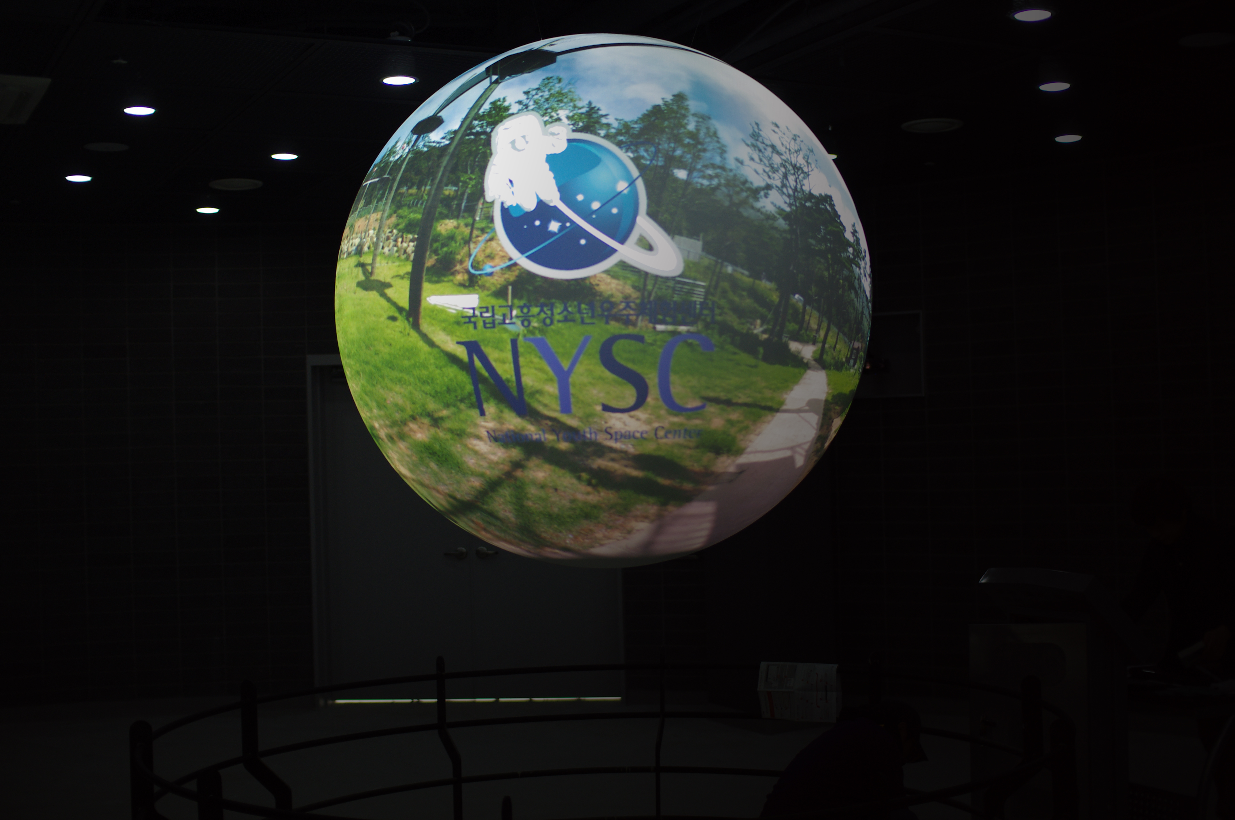 Science On a Sphere displays the National Youth Space Center logo atop a photograph including a sidewalk, some grass, and some trees