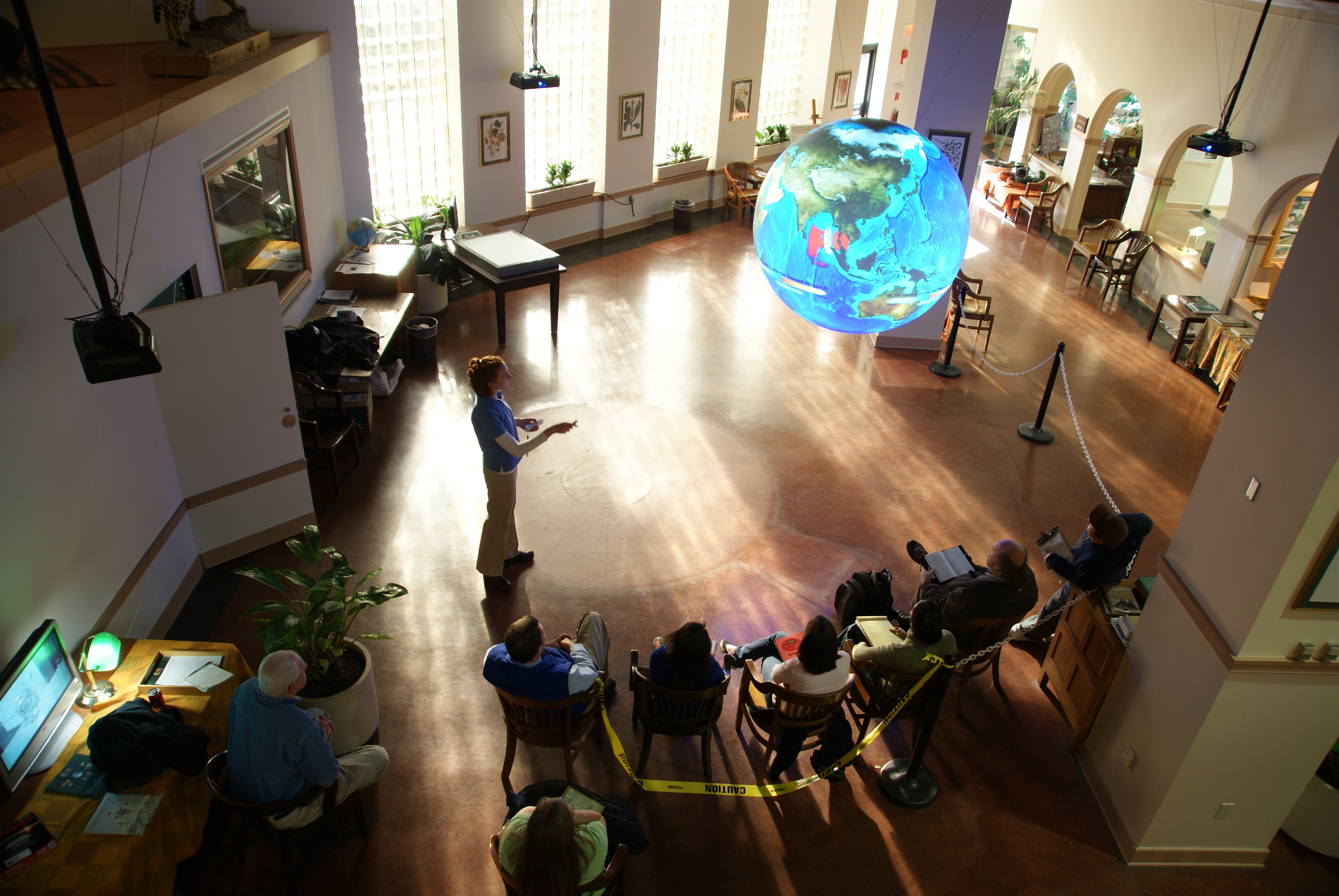 The camera looks down on a group of adults watching a presentation on Science On a Sphere. Behind the Sphere are a set of tall windows, the light from which casts long shadows on the floor