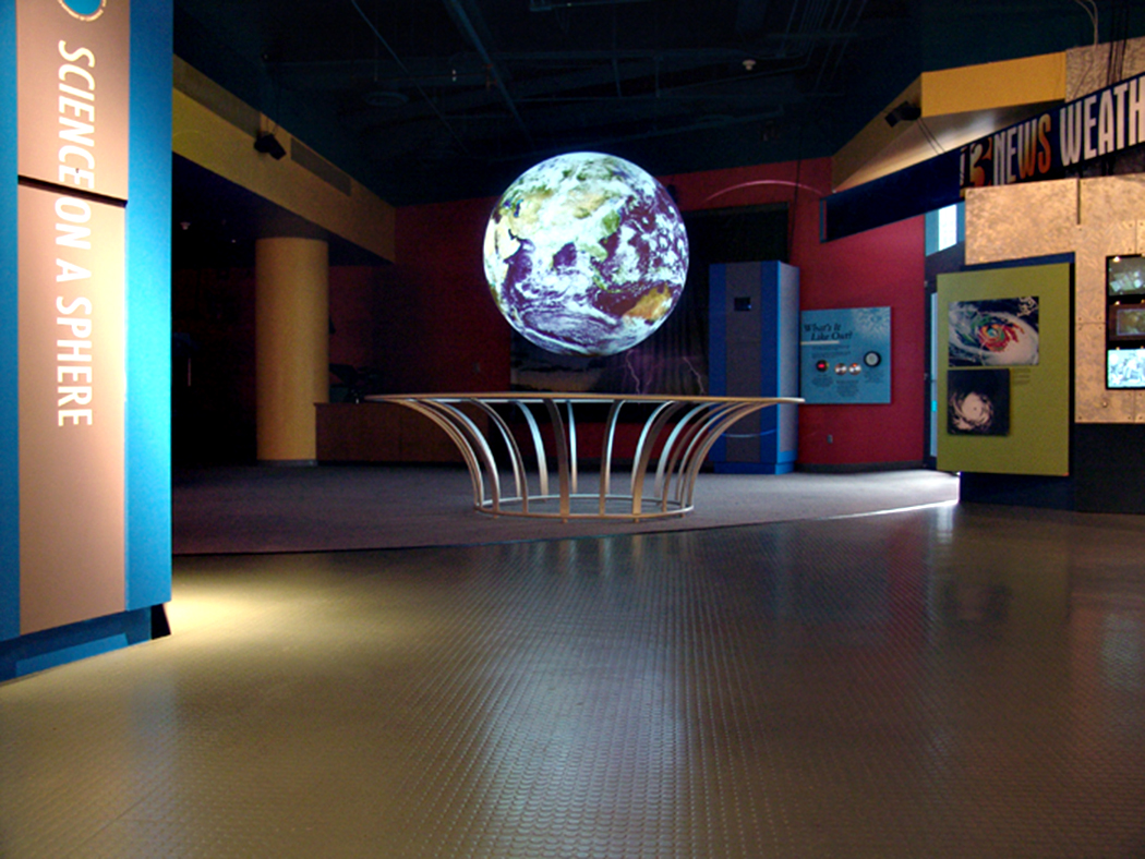 Science On a Sphere hangs in an empty exhibity space. Two images of hurricanes are mounted on the wall to the right