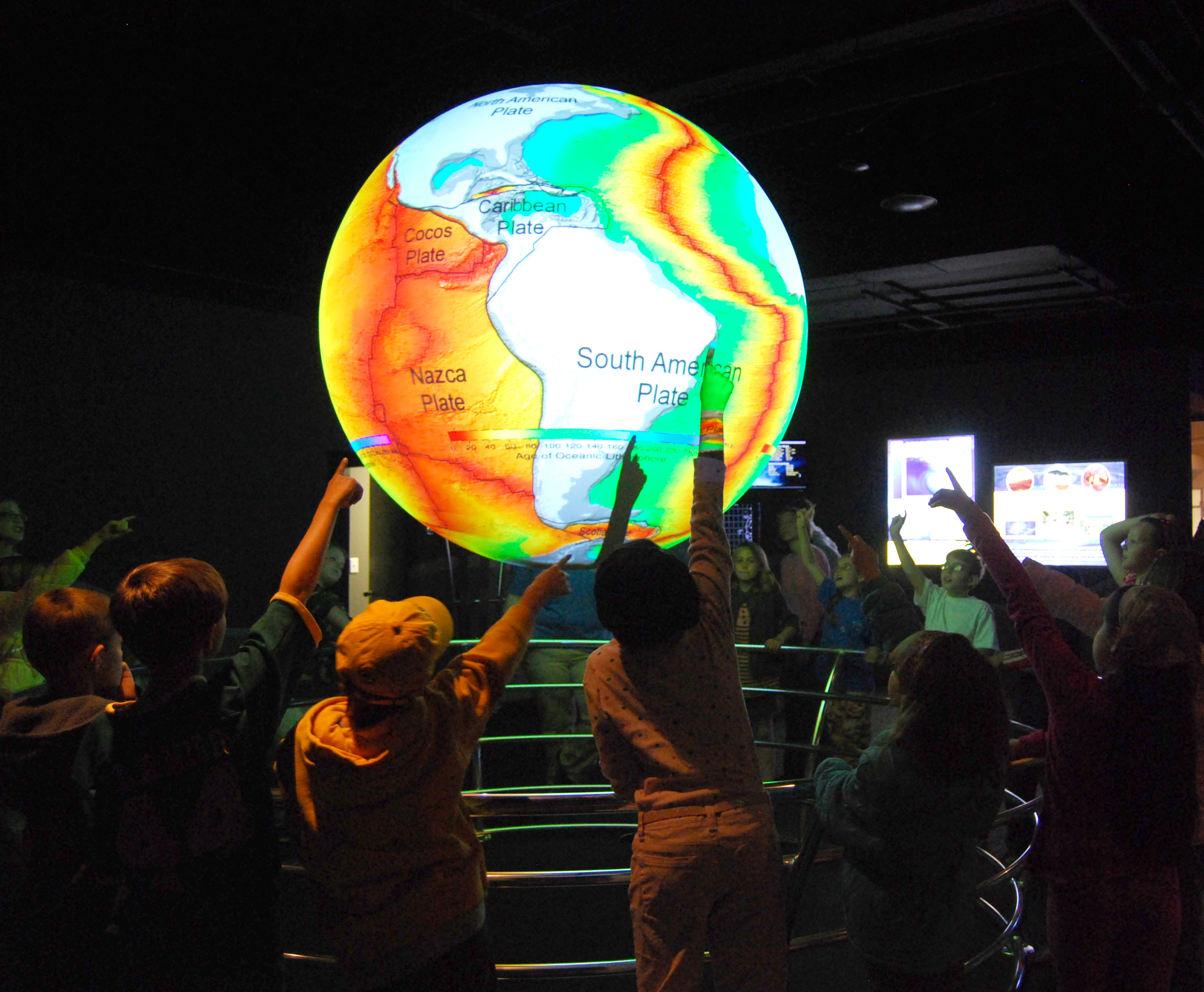 A group of children circle Science On a Sphere, pointing at it. The Sphere displays the age of the sea floor in hues of red, yellow, and green