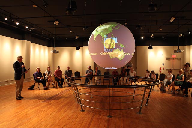 A group of people sit on chairs in the background watching a presentation on Science On a Sphere in a well-lit theater. The Sphere displays the logo for the Nurture Nature center of an image of the Earth