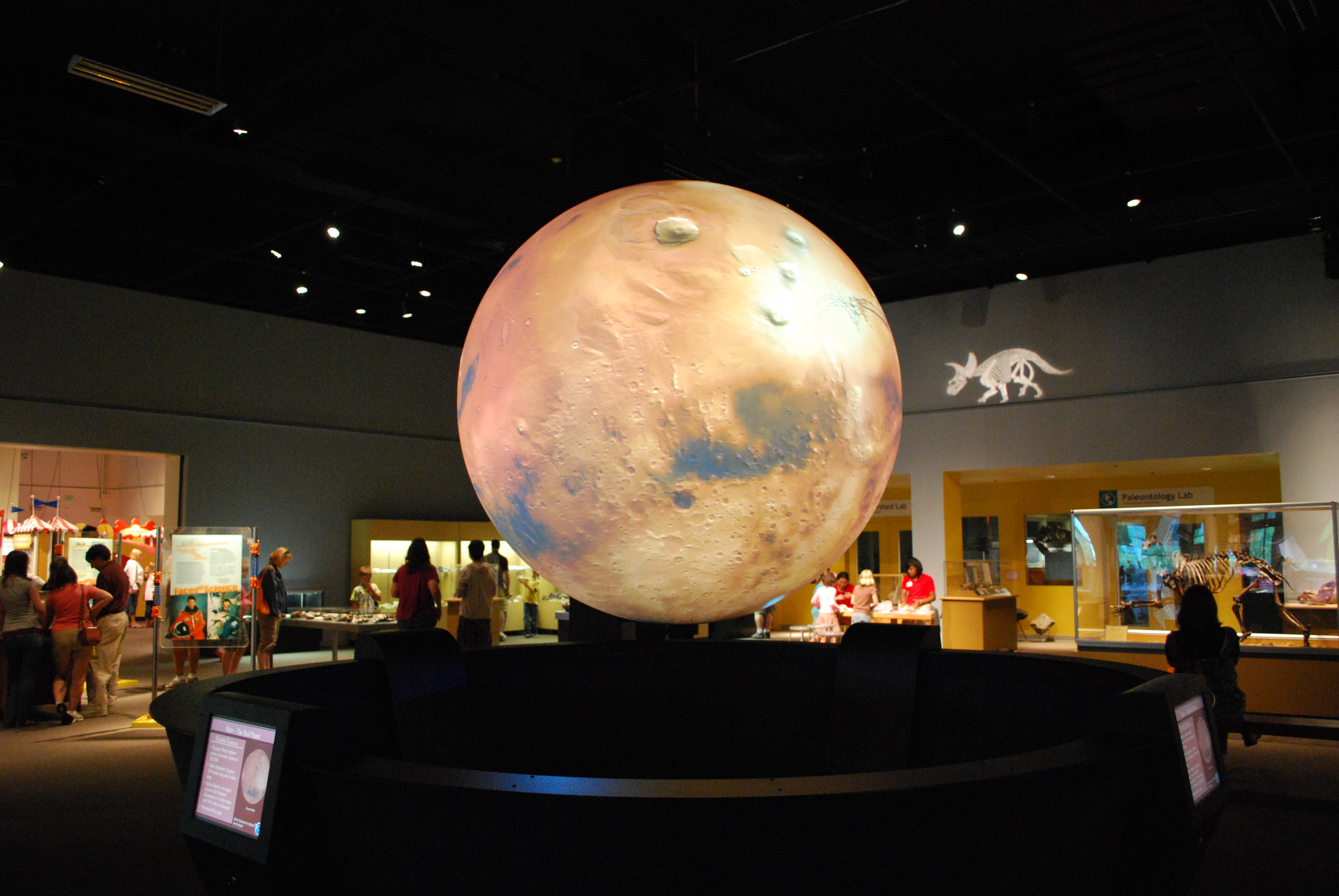 Science On a Sphere hangs in an exhibit hall displaying an image of Earth's moon. In the background, museum-goers explore other exhibits at the museum