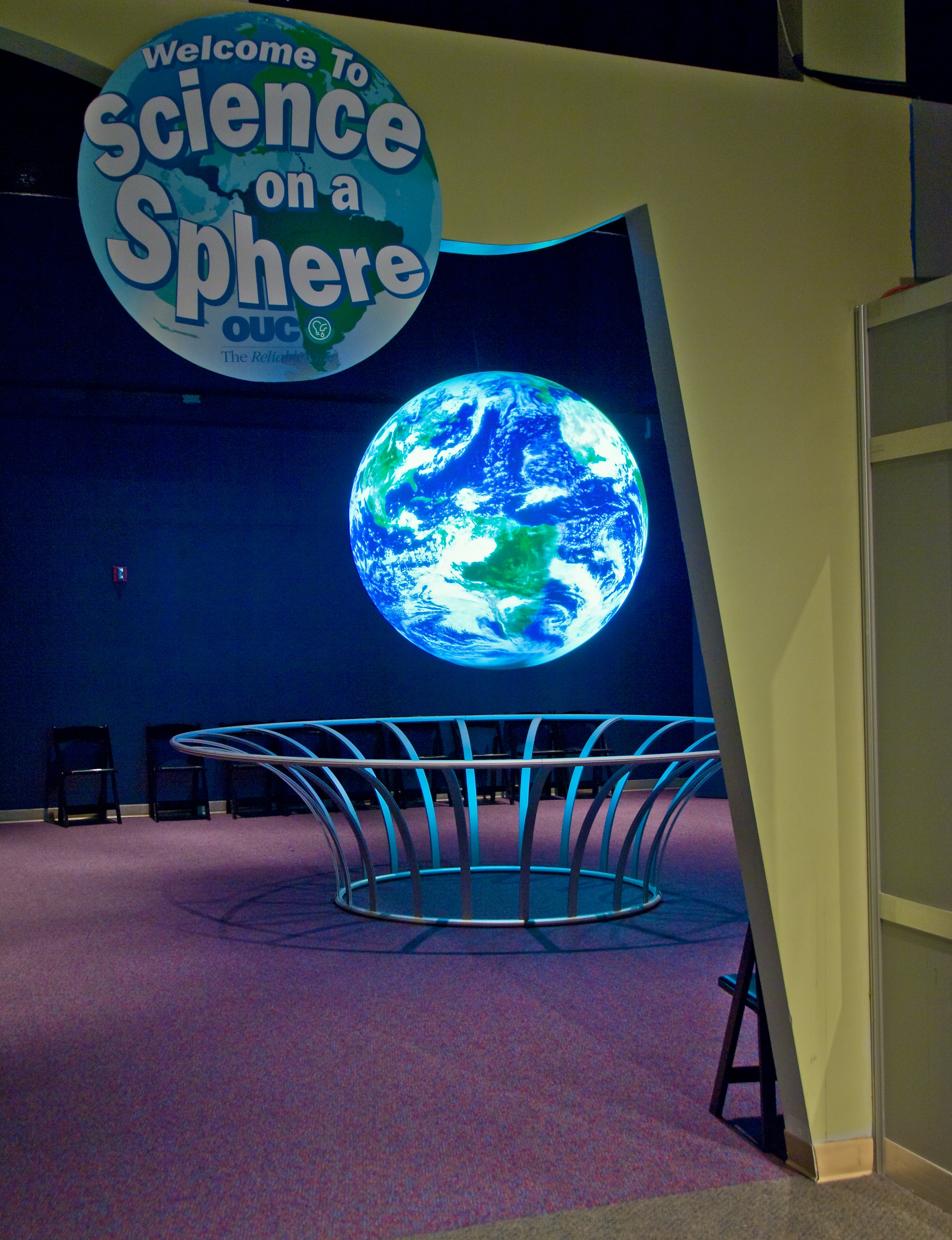 Science On a Sphere is visible through an entryway beneath a sign that reads 'Welcome to Science On a Sphere'. Several folding chairs line the wall behind the Sphere