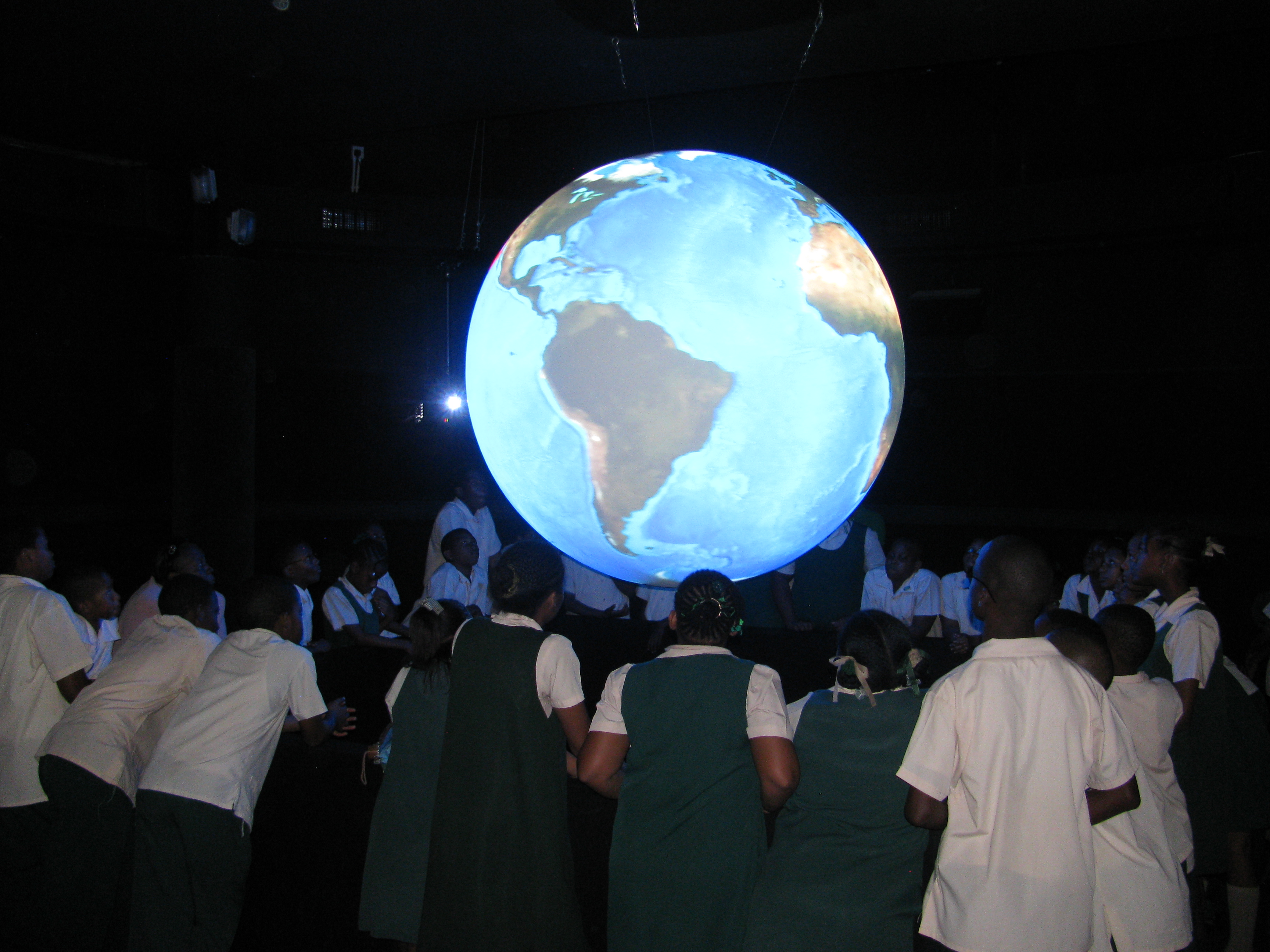 A group of students in uniforms stand in a circle around Science On a Sphere in a darkened room