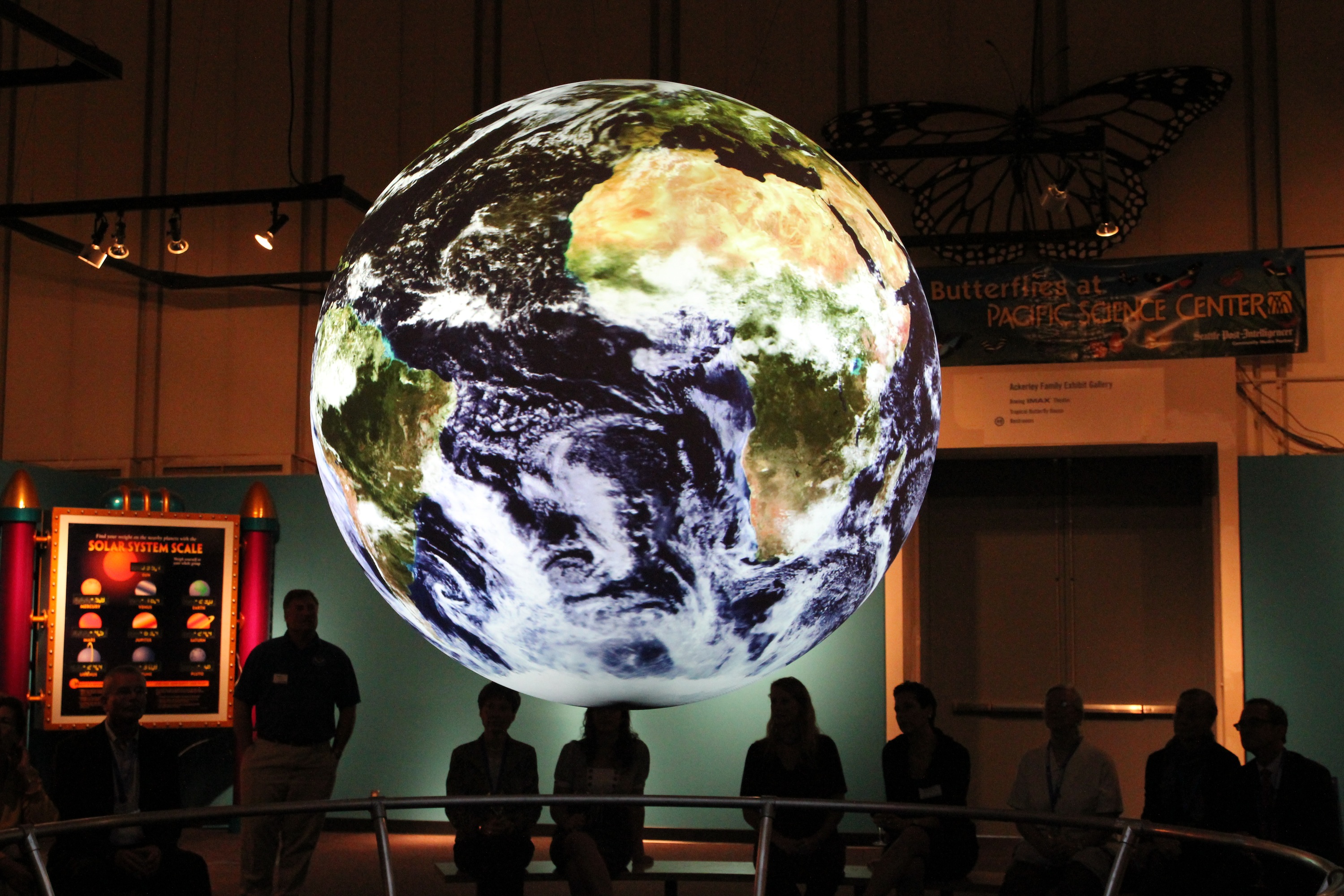 Science On a Sphere displays satellite imagery of Earth, behind the Sphere can be seen silhouettes of people sitting on benches watching the Sphere
