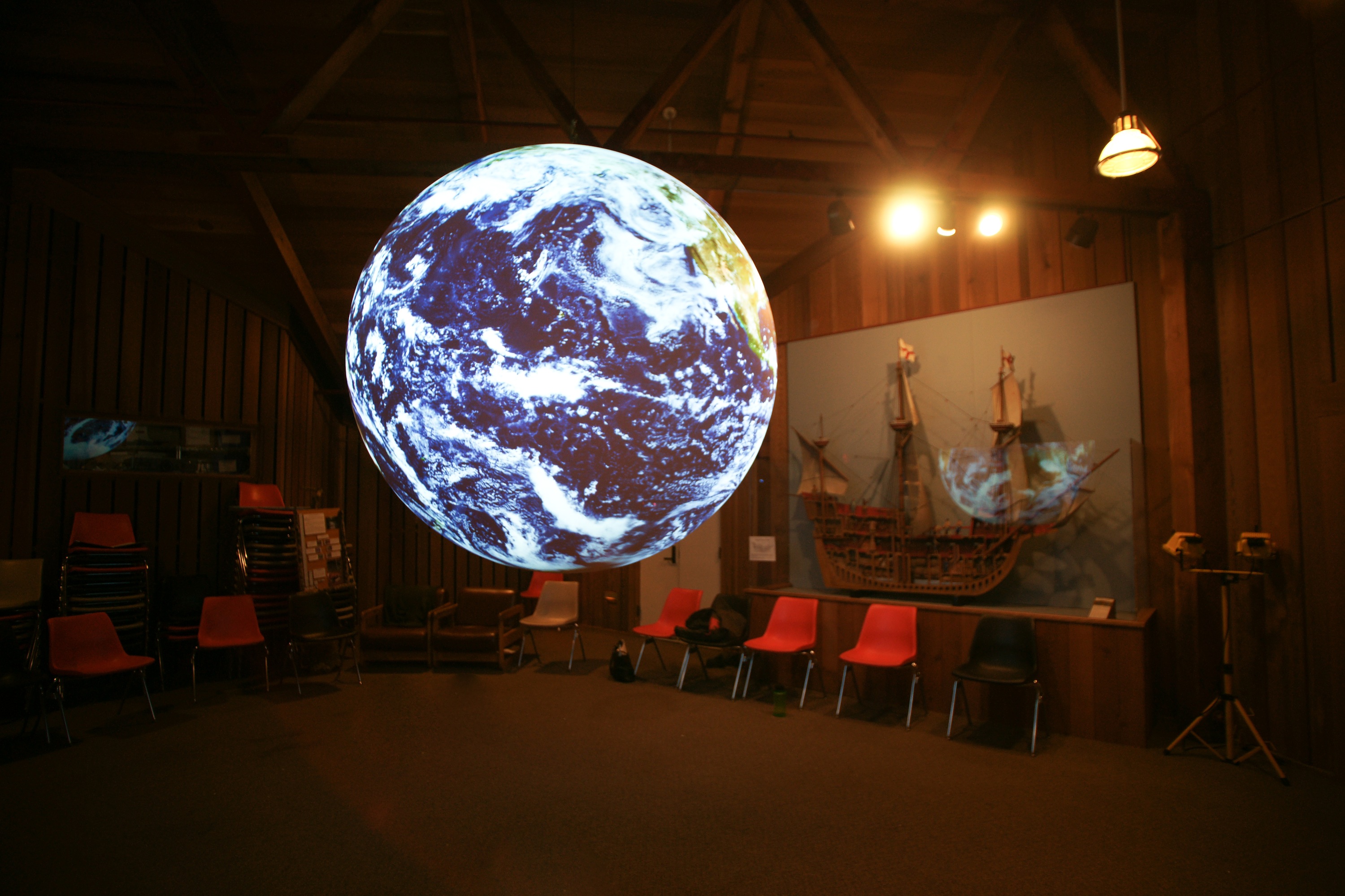 Science On a Sphere displayse satellite imagery of the Earth in a room with wooden walls. In the background the celing rafters are visible. Behind the Sphere sit stacks of chairs and a cross-section of an old sailing ship