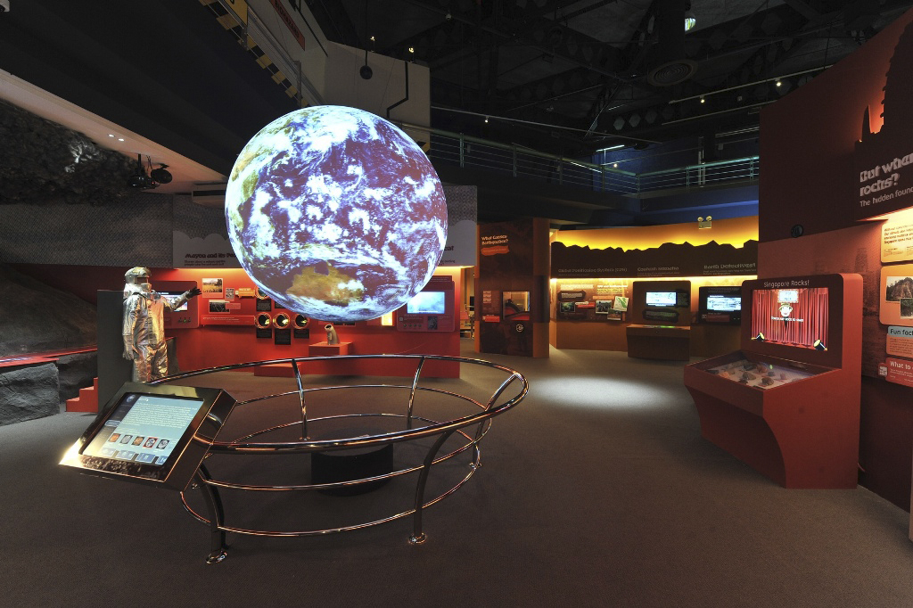 Science On a Sphere hangs in an empty exhibit space. In the background other exhibits relating to geology are visible