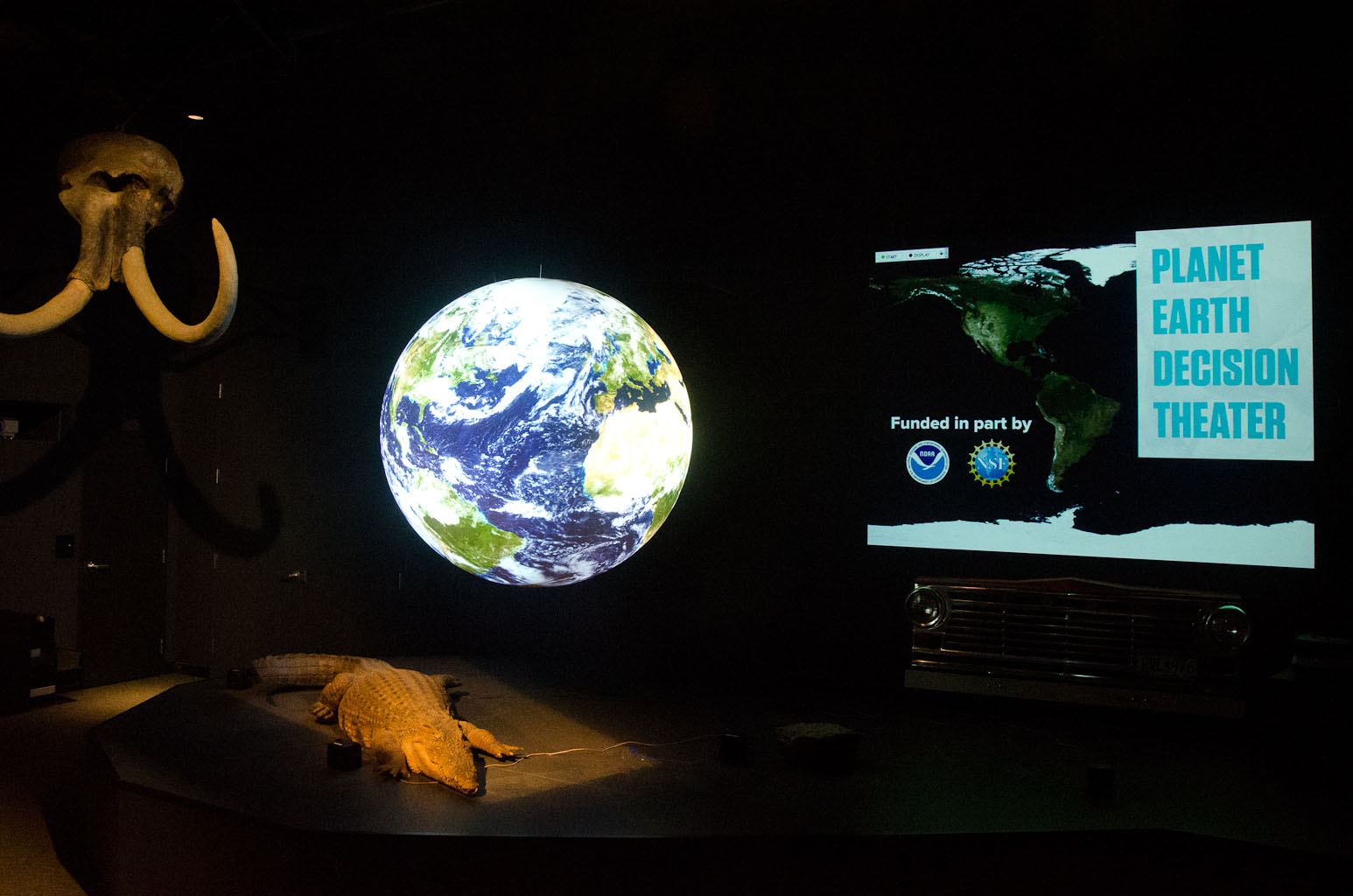 Science On a Sphere hangs in a darkened room. To the left is an elephant skull, and beneath the Sphere is a crocodile. To the right is a digital display reading 'Planet Earth Decision Theater'