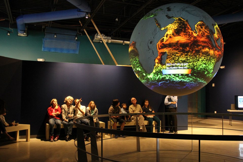 A group of visitors sit on benches watching Science On a Sphere as it displays colorful, swirling lines in hues of red, yellow, green, and blue in the ocean