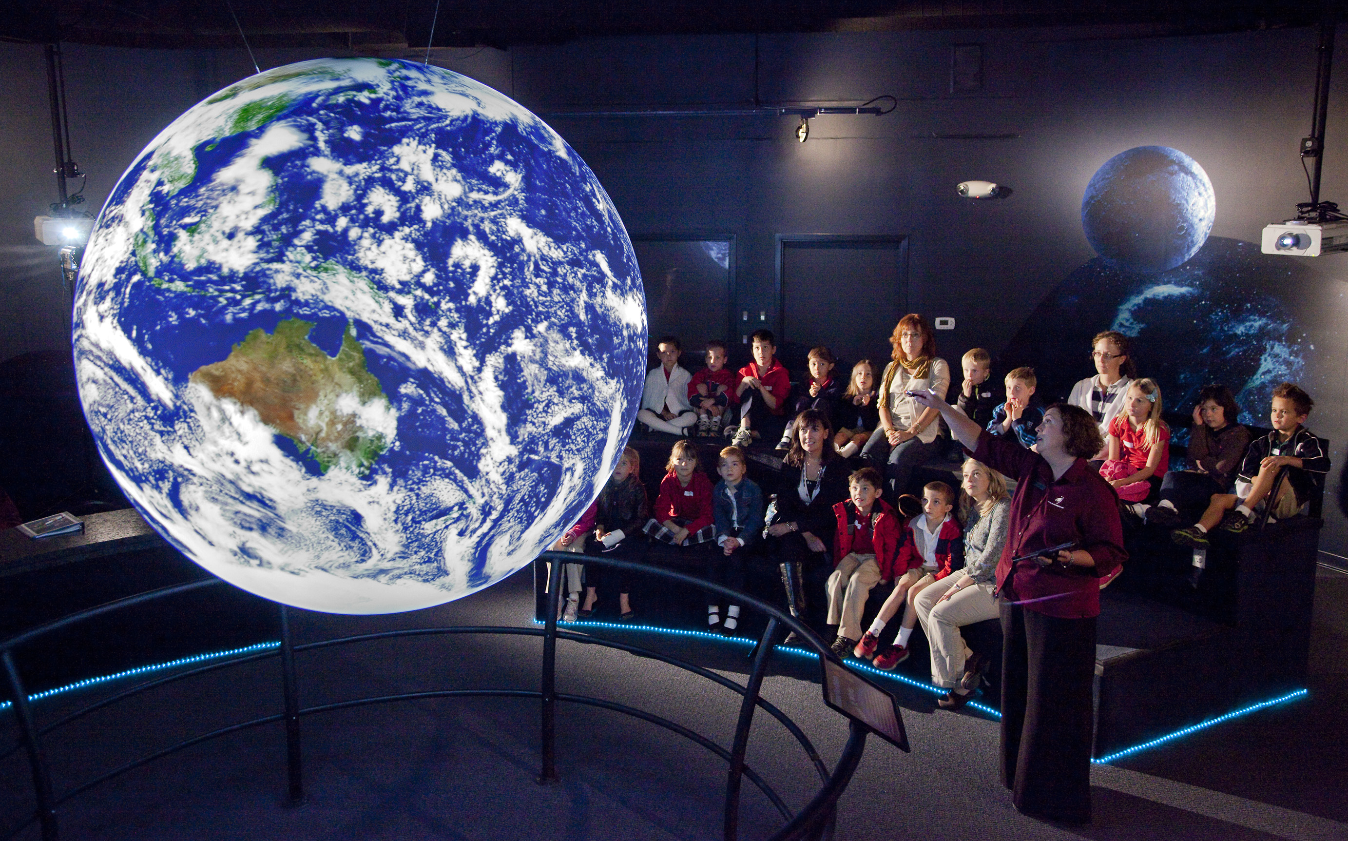A group of school children sit on a set of terraced benches watching a presentation by a docent on Science On a Sphere