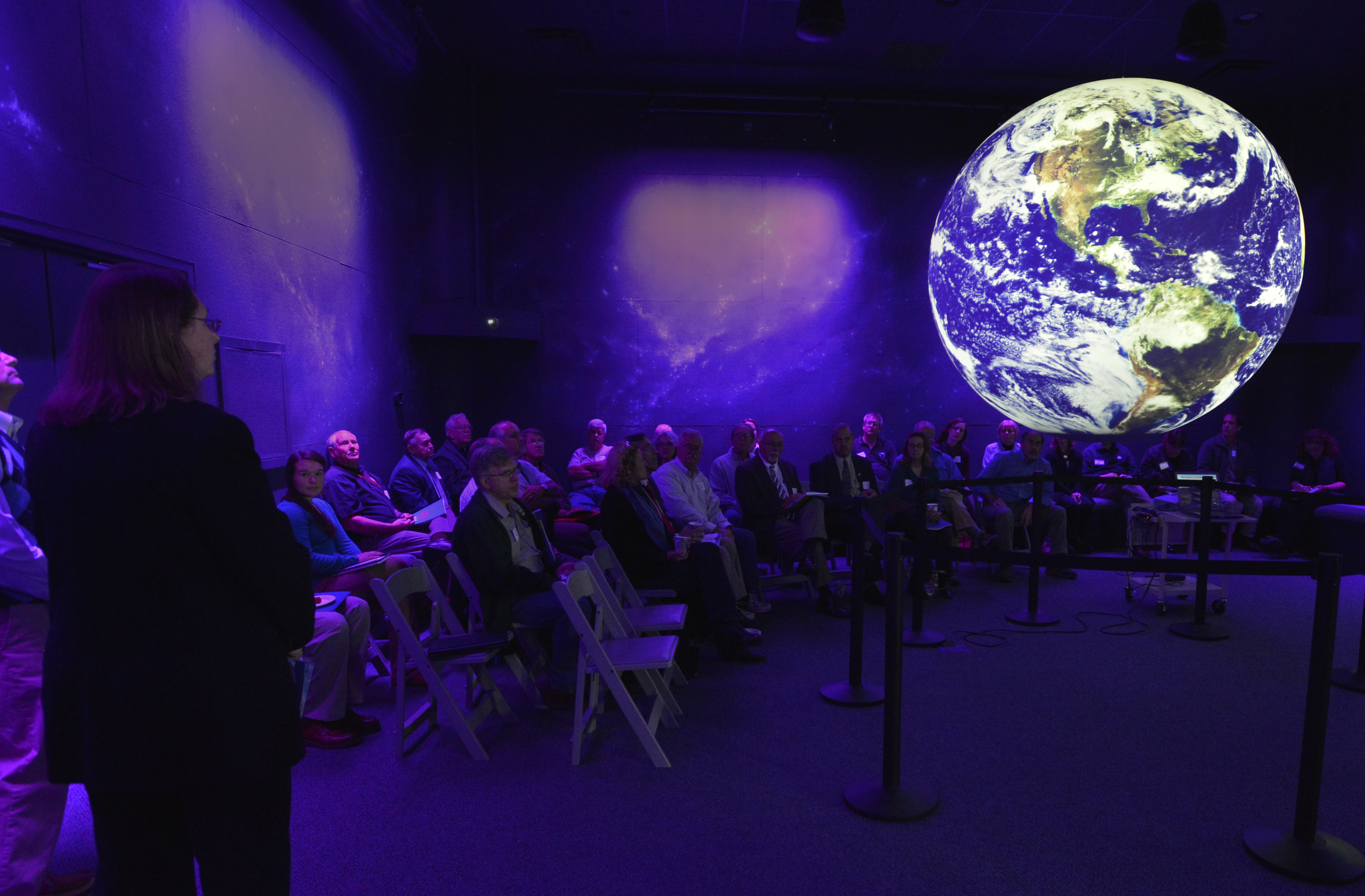 An audience sits on folding chairs in a quarter circle around Science On a Sphere. The lighting in the room creates a purplish cast to everything except the Sphere, which is a bright blue and white as it displays satellite imagery of Earth
