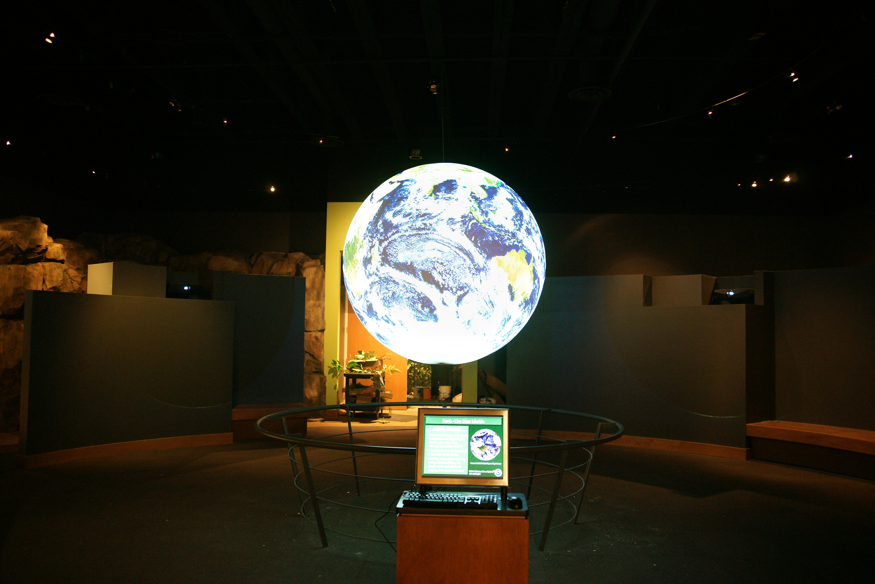 Science On a Sphere displays satellite imagery of Earth in an empty room. In front of the Sphere sits a computer monitor, keyboard, and mouse