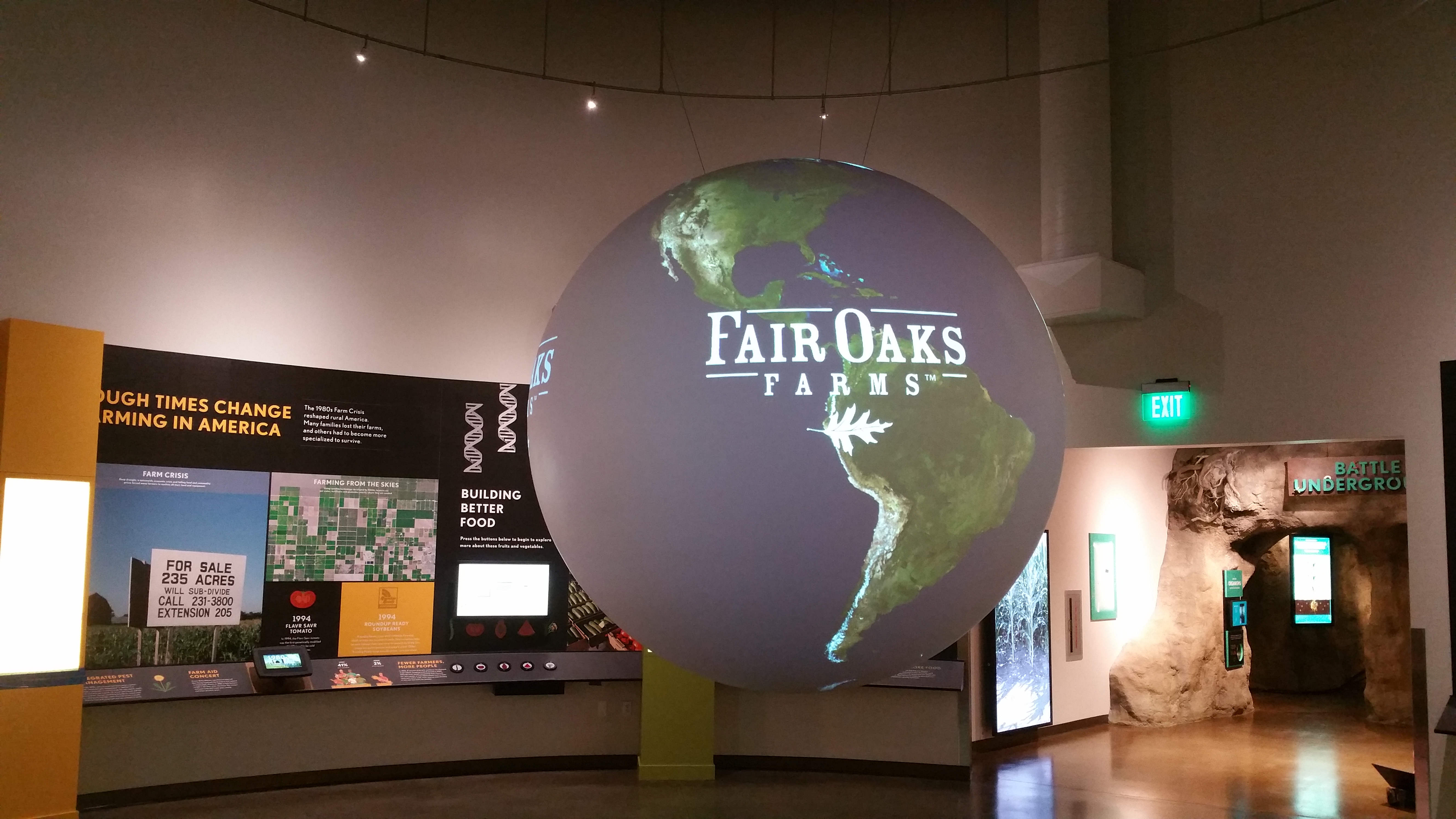 Science On a Sphere displays the Fair Oaks Farm logo over an image of Earth in a well-lit room. Behind it can be seen an exhibit on farming in America