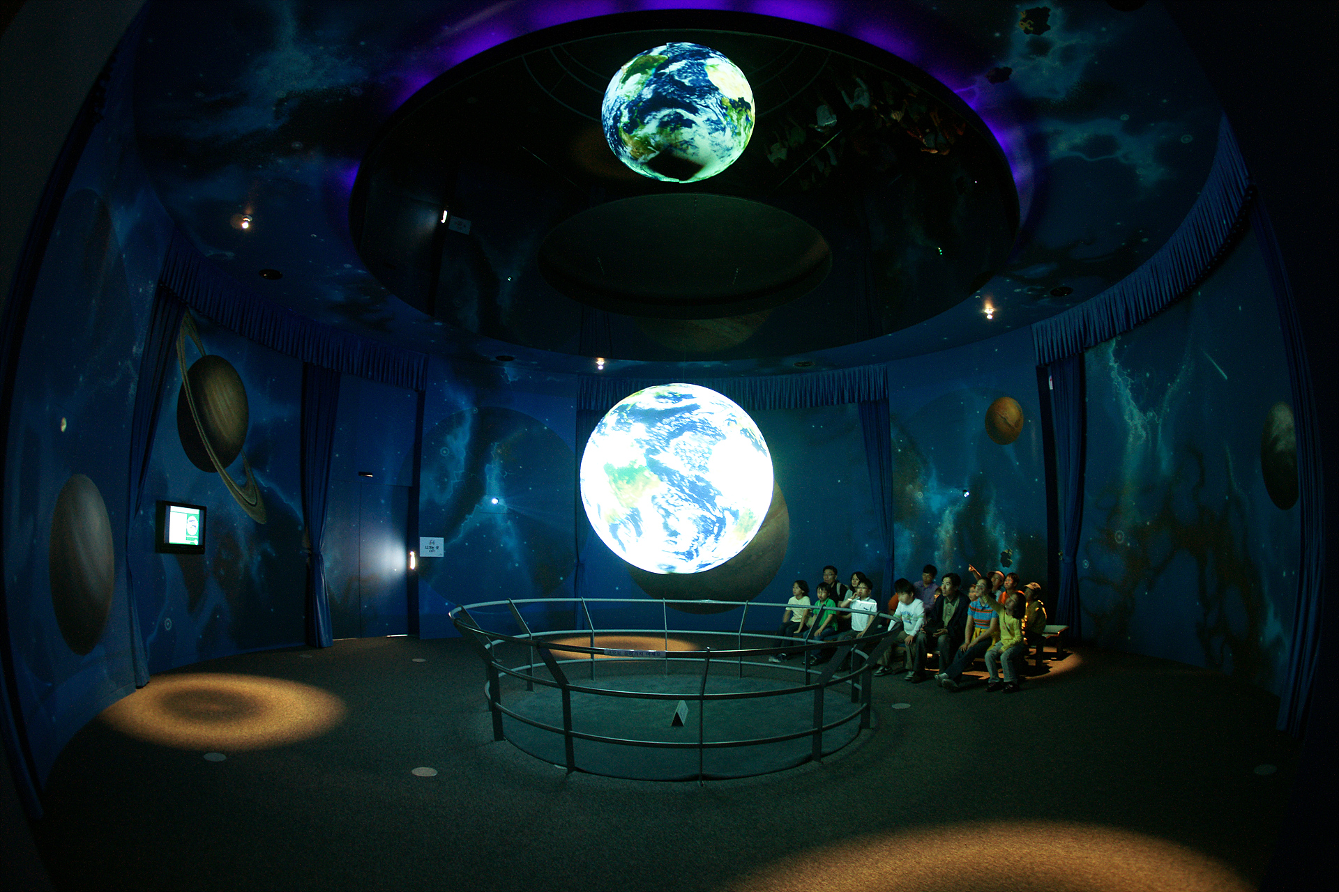 A group of people sit on benches to one side in a room watching Science On a Sphere. The walls of the room are painted with images of planets and nebulae. The Sphere is reflected in the ceiling by a fixture surrounding where it's mounted