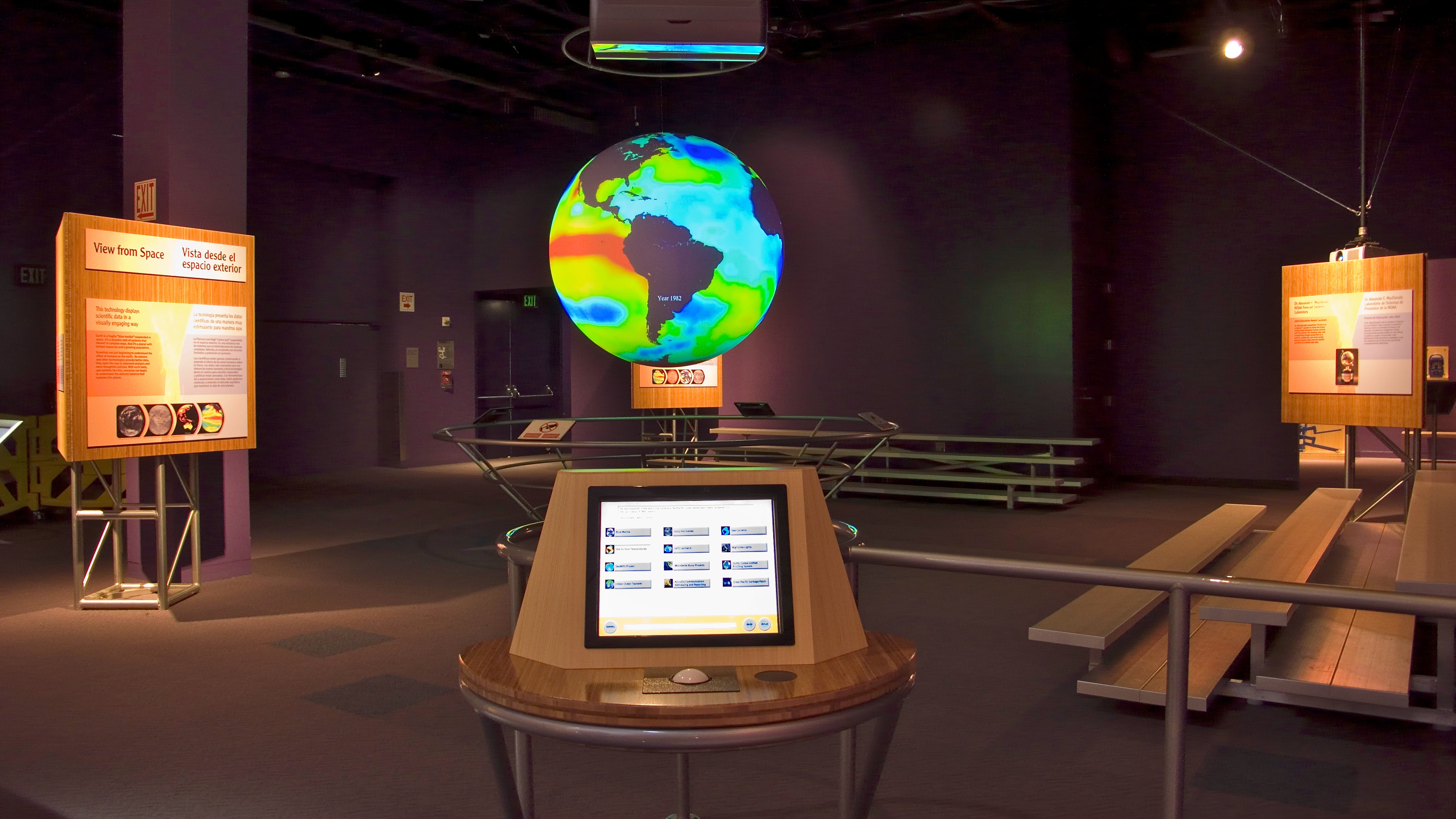 Science On a Sphere hangs in an empty exhibit space. A monitor with a track ball sits on a table in front of the Sphere. Two sets of risers are situated around the Sphere, and placards containing additional information are visible in the background